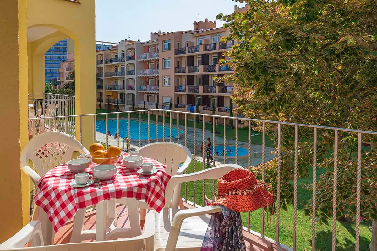 Terraza soleada con mesa, desayuno y vista a piscina y edificios.