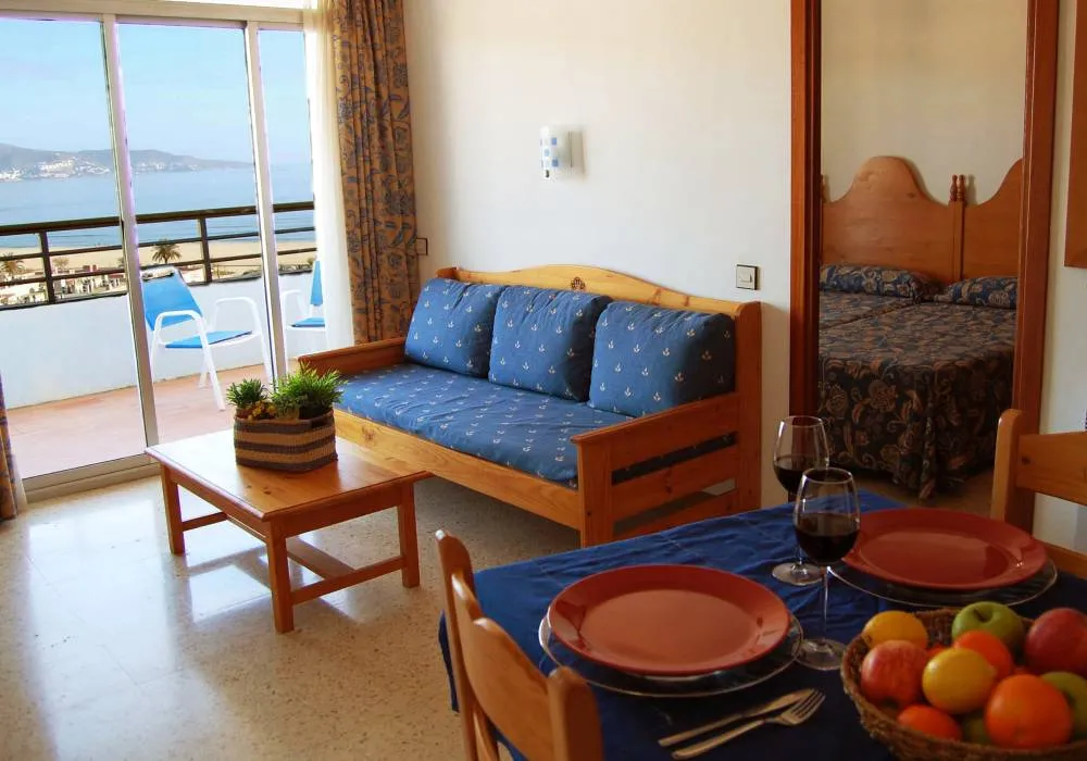 Sala luminosa con vista al mar, sof&aacute; azul, mesa con fruta y dormitorio.