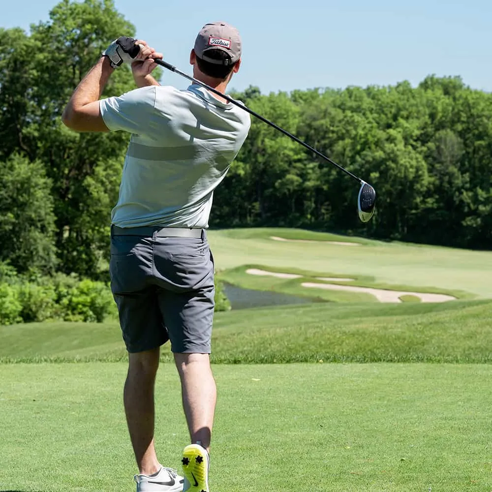 Persona jugando golf en un campo verde, con &aacute;rboles al fondo, d&iacute;a soleado.