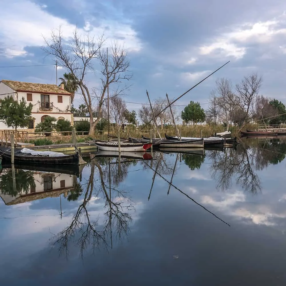 Barcas junto a una casa al borde de un lago, reflejadas en el agua tranquila.