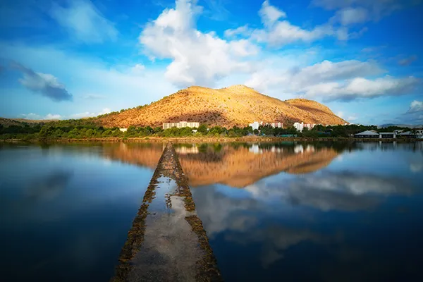 Paisaje con monta&ntilde;a reflejada en el agua bajo un cielo azul con nubes.