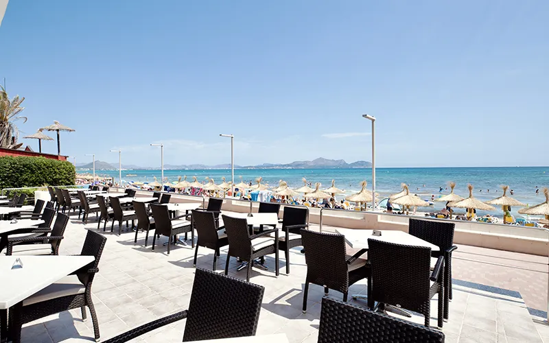 Terraza junto al mar con sillas negras y playa llena de sombrillas y ba&ntilde;istas.