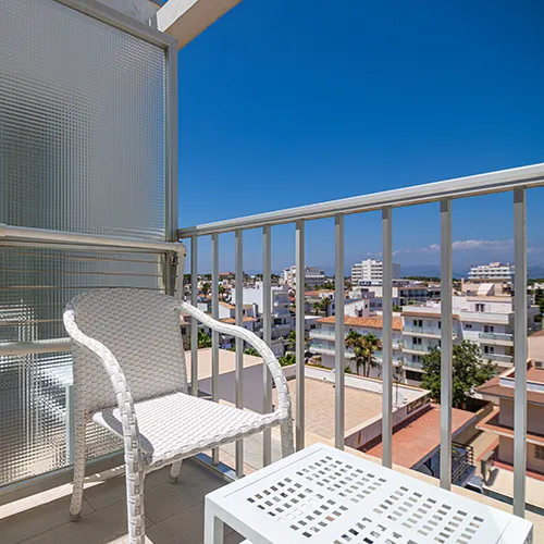 Balc&oacute;n con silla blanca, mesa y vista urbana bajo un cielo azul despejado.