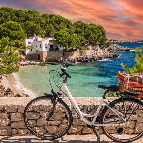 Bicicleta frente a una playa y casa rodeada de &aacute;rboles bajo un cielo colorido.