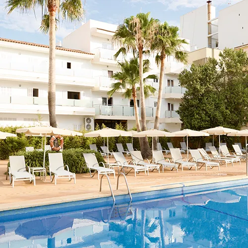 Piscina con tumbonas y sombrillas junto a un edificio blanco rodeado de palmeras.