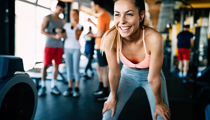 Mujer sonriendo en el gimnasio, personas desenfocadas al fondo.