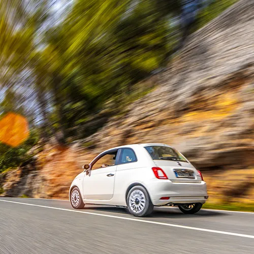 Auto blanco conduciendo r&aacute;pido en una carretera con fondo borroso de &aacute;rboles y rocas.