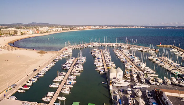Puerto deportivo junto a una playa de arena con edificios y monta&ntilde;as al fondo.