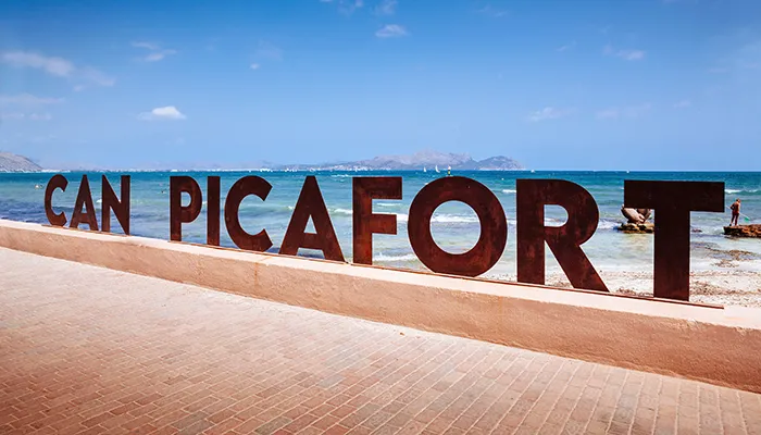 Letrero de Can Picafort con el mar de fondo en un d&iacute;a soleado.
