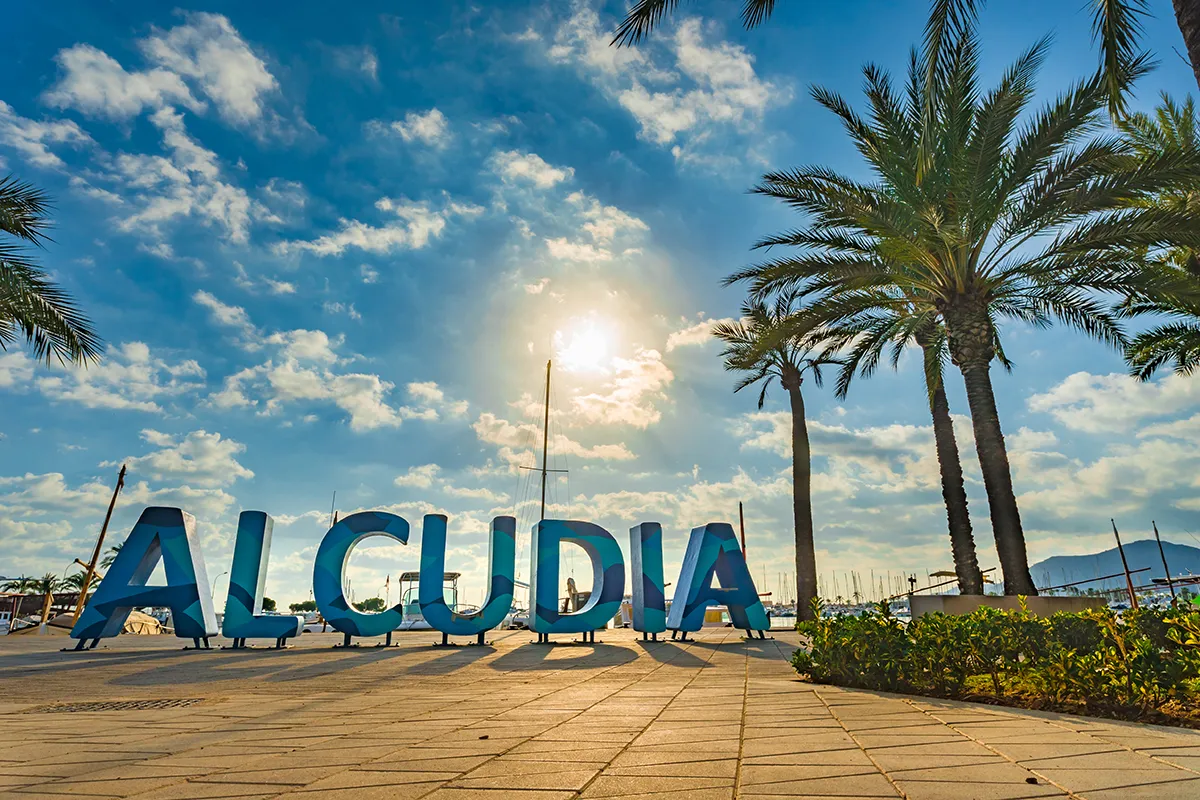 Letras "ALCUDIA" con palmeras y cielo azul al fondo.