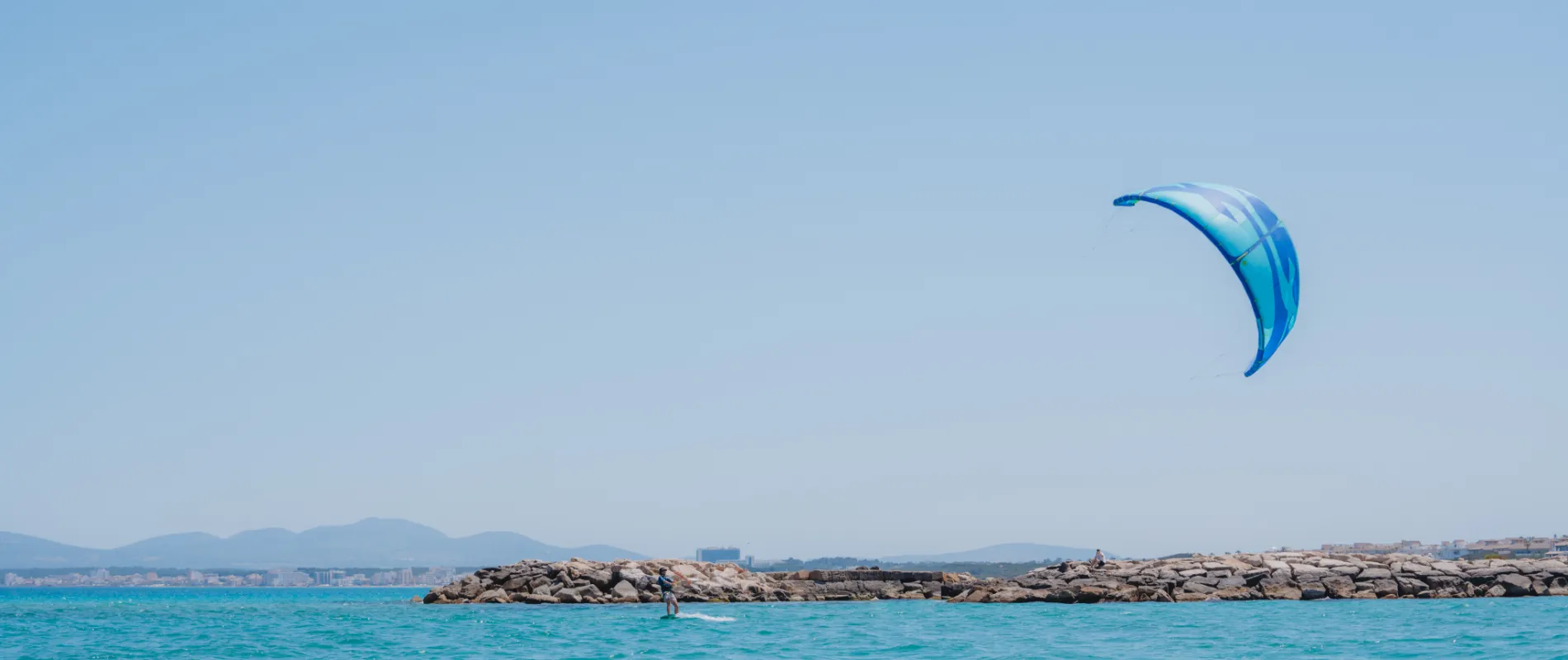 Surfista con cometa azul en el mar, junto a un rompeolas.