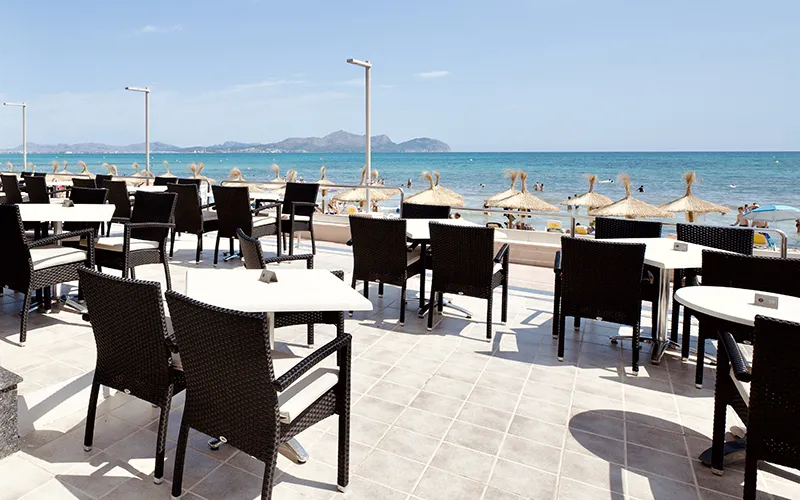 Terraza vac&iacute;a con mesas, frente a la playa y el mar azul bajo un cielo despejado.