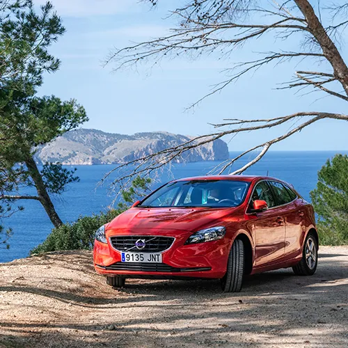 Coche rojo estacionado frente al mar con monta&ntilde;as al fondo.