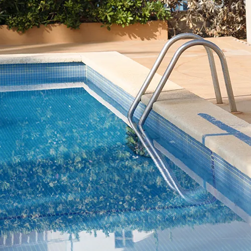 Escalera met&aacute;lica en una piscina azul con borde de baldosas blancas y marrones.