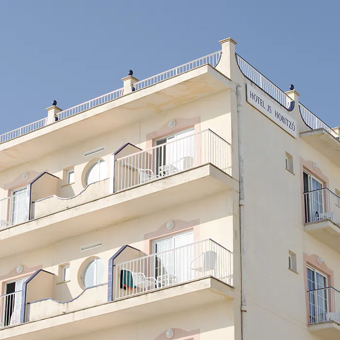 Fachada clara de un hotel con barandillas y ventanas al sol.