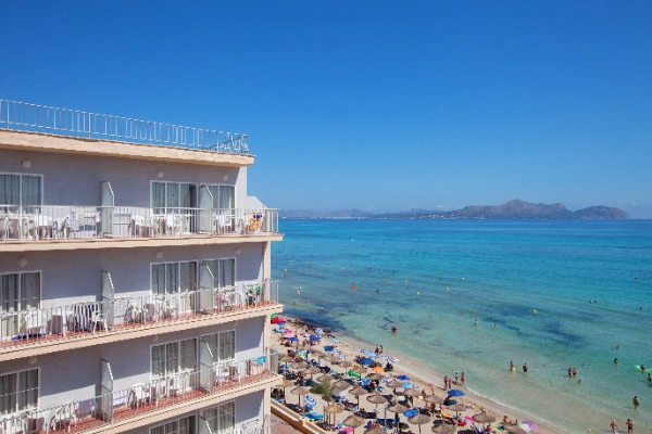 Edificio junto a una playa concurrida con mar azul y sombrillas coloridas.