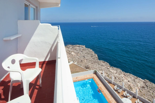 Balc&oacute;n con silla blanca, vista al mar y piscina junto a un acantilado rocoso.