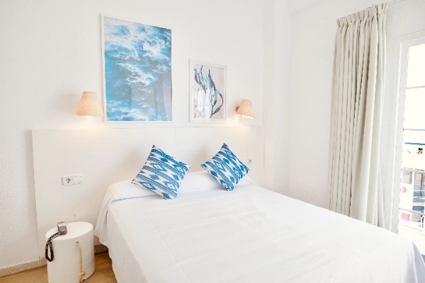 Habitaci&oacute;n minimalista con cama, cojines azules, l&aacute;mparas y cuadros en la pared.