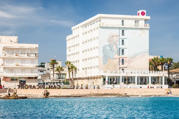 Hotel junto a la playa con un mural en una pared, cielo azul y palmeras.