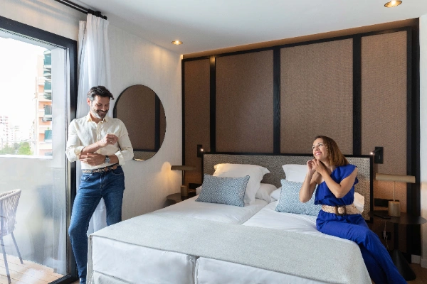 Pareja en habitaci&oacute;n de hotel, &eacute;l sonr&iacute;e y ella est&aacute; sentada en la cama.