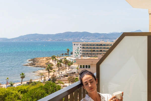 Mujer tomando caf&eacute; en un balc&oacute;n con vista al mar y colinas al fondo.