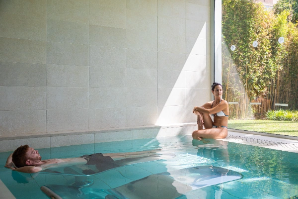 Dos personas relaj&aacute;ndose en una piscina cubierta junto a un ventanal.