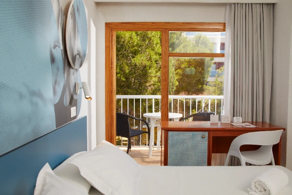 Habitaci&oacute;n luminosa con balc&oacute;n, mesa y sillas, vista a &aacute;rboles.