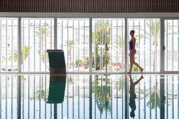 Persona en traje de ba&ntilde;o junto a una piscina interior con ventanas y palmeras afuera.