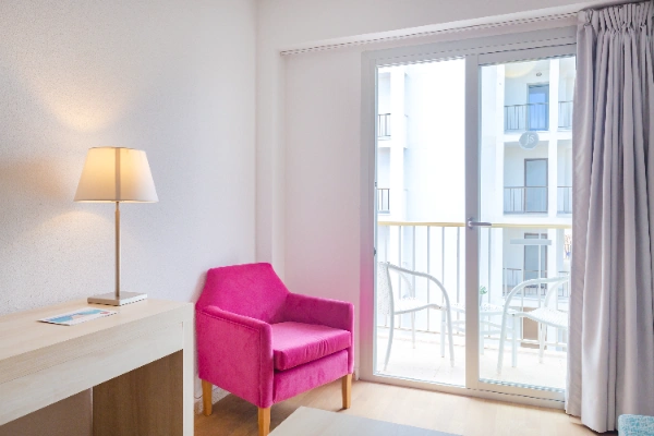 Habitaci&oacute;n con sill&oacute;n rosa, l&aacute;mpara y puerta ventana hacia un balc&oacute;n.