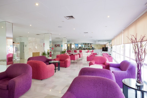 Sala de espera con sof&aacute;s morados y mesas, iluminada por ventanas grandes.