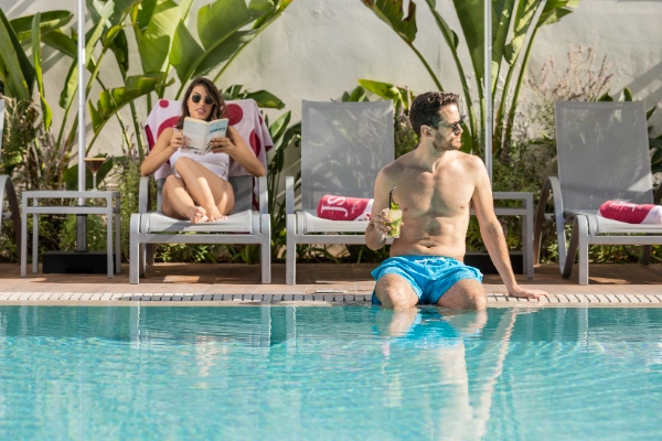 Hombre en piscina con bebida, mujer leyendo tras &eacute;l, rodeados de vegetaci&oacute;n.