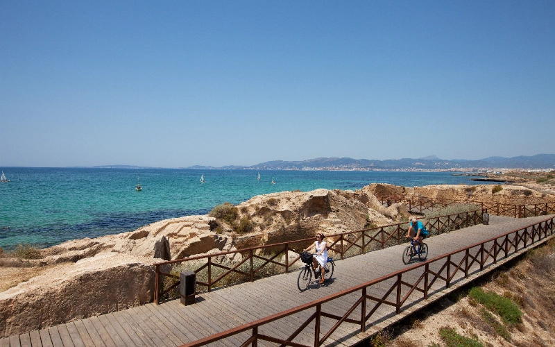 Dos personas en bicicleta por un paseo costero junto al mar azul.