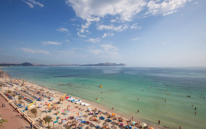 Playa concurrida con sombrillas y ba&ntilde;istas, mar turquesa y cielo parcialmente nublado.