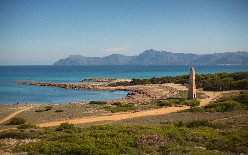 Paisaje costero con obelisco, mar azul y monta&ntilde;as al fondo.