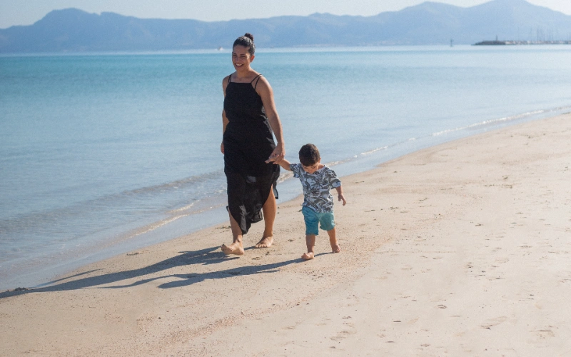 Mujer y ni&ntilde;o caminando descalzos en la playa junto al mar azul.