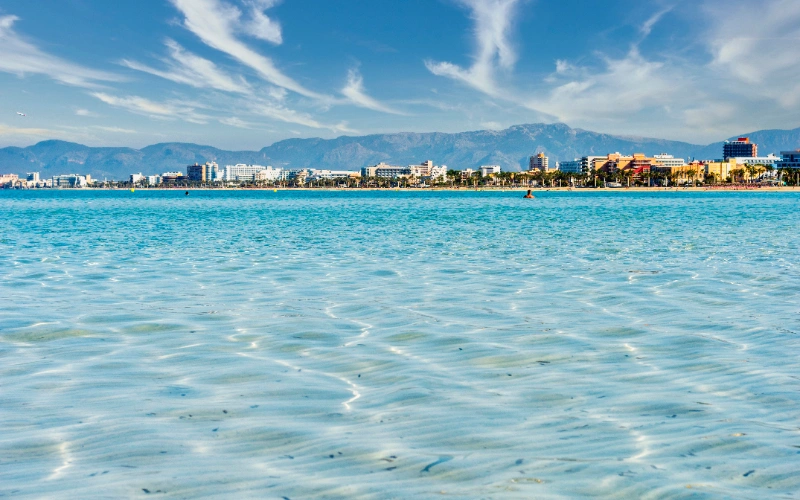 Agua cristalina, ciudad y monta&ntilde;as bajo un cielo azul y nubes.