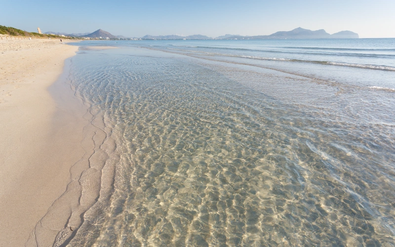 Playa con arena clara y agua cristalina, monta&ntilde;as al fondo.
