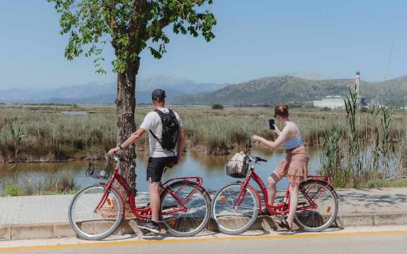 Personas con bicicletas junto a un r&iacute;o, paisaje monta&ntilde;oso al fondo.