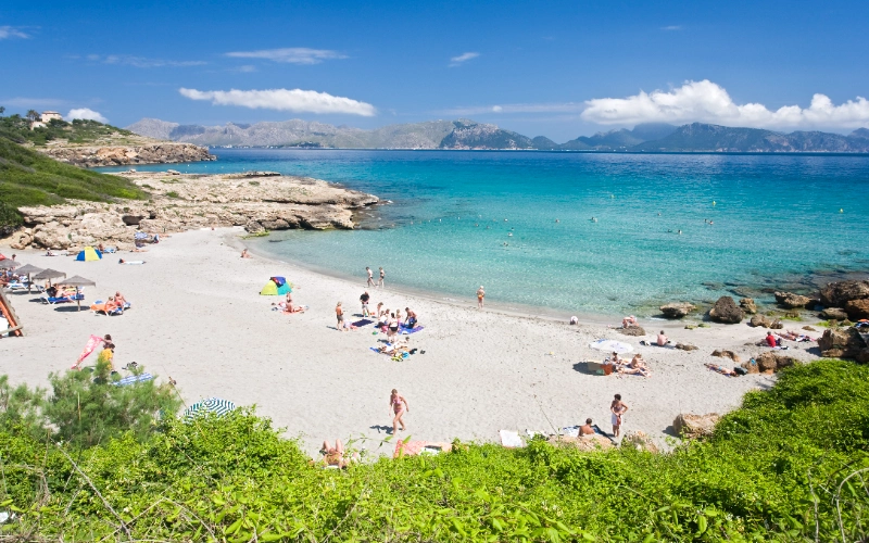 Playa con arena blanca, personas y sombrillas; mar azul y monta&ntilde;as al fondo.