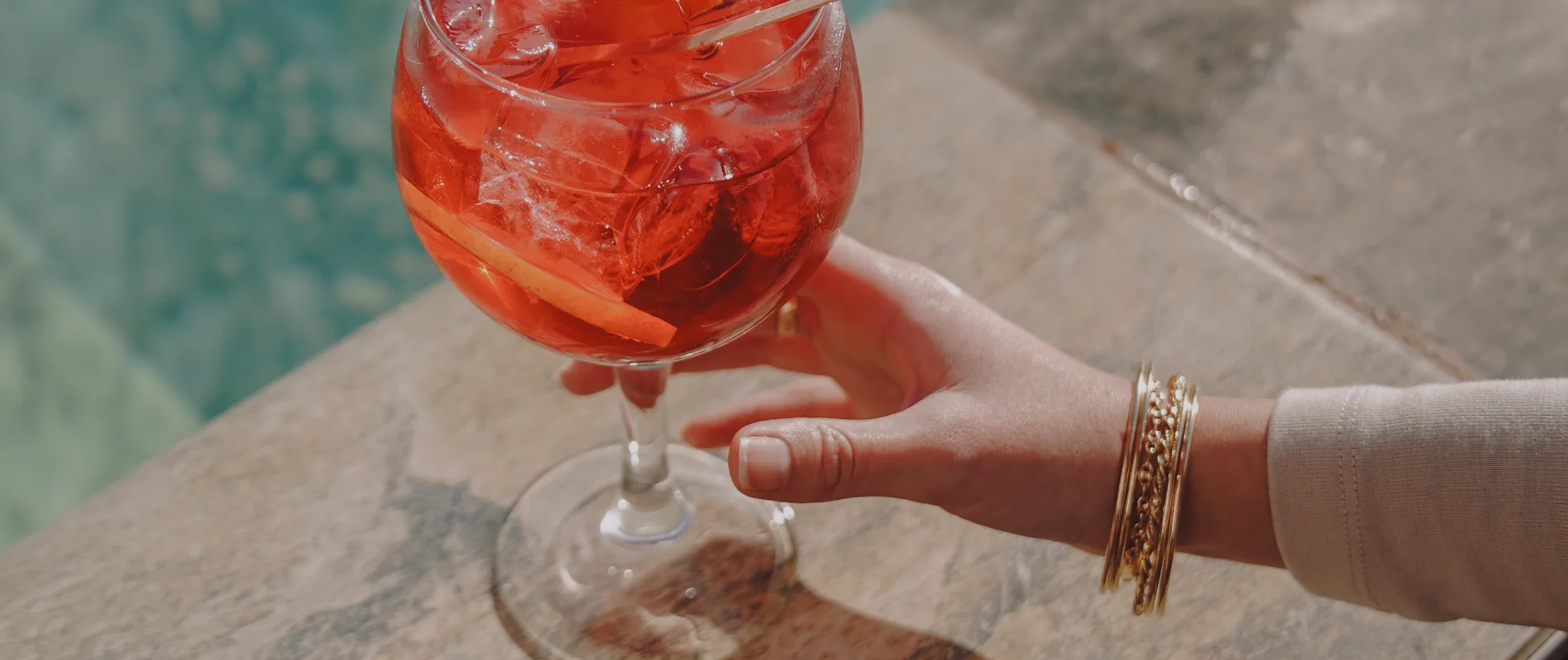 Mano sujetando copa con bebida roja y hielo, junto a piscina.