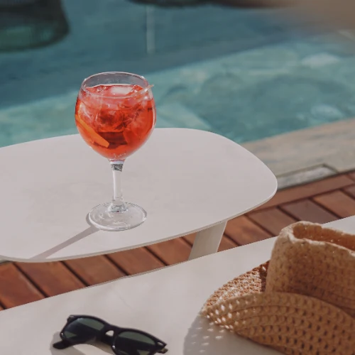 Copa con bebida junto a una piscina, sombrero y gafas de sol sobre la mesa.