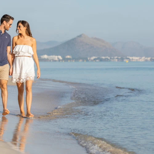 Pareja caminando por la playa en un d&iacute;a soleado con monta&ntilde;as al fondo.