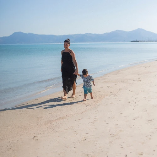 Mujer y ni&ntilde;o caminando en la playa con el mar y monta&ntilde;as al fondo.