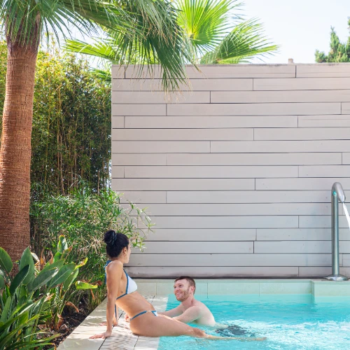 Persona sentada y otra en la piscina, rodeadas de palmeras y vegetaci&oacute;n.