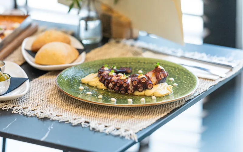 Plato de pulpo a la parrilla con pur&eacute; y pan, sobre una mesa elegante.