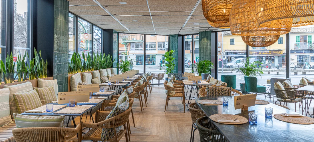 Restaurante moderno con luz natural, mesas de mimbre y decoraci&oacute;n vegetal.