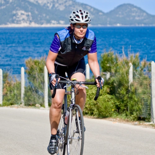 Persona en bicicleta junto al mar, con colinas de fondo.