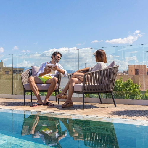 Dos personas conversando junto a una piscina en una terraza soleada.