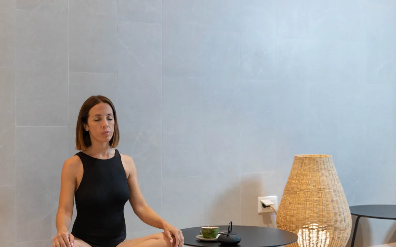 Mujer meditando en posici&oacute;n de loto junto a mesa con taza y l&aacute;mpara de mimbre.