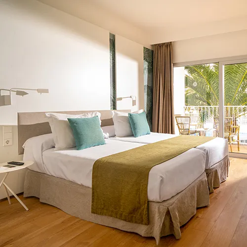 Habitaci&oacute;n de hotel con dos camas, decoraci&oacute;n c&aacute;lida y ventanal hacia el balc&oacute;n.